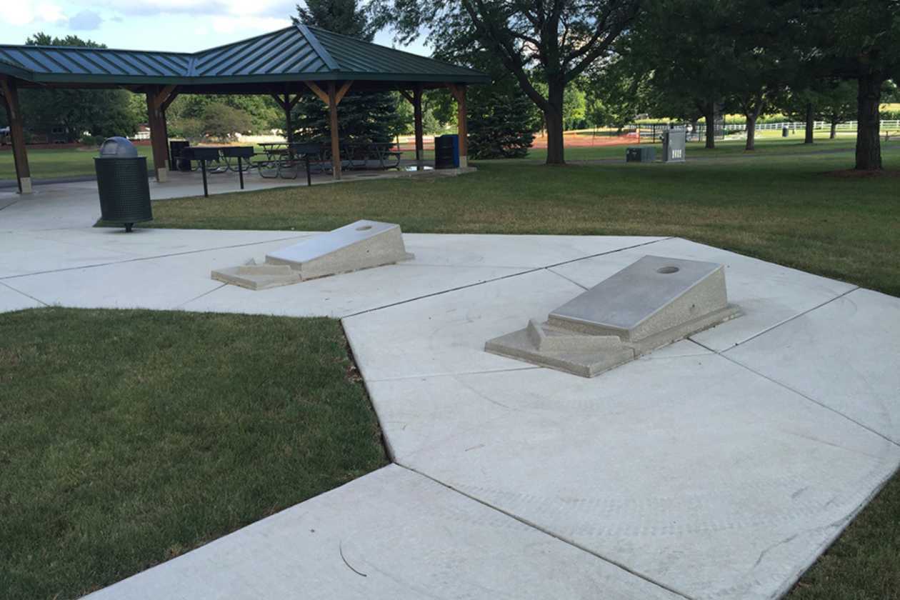 Concrete cornhole boards on a paved recreation area near a covered picnic space